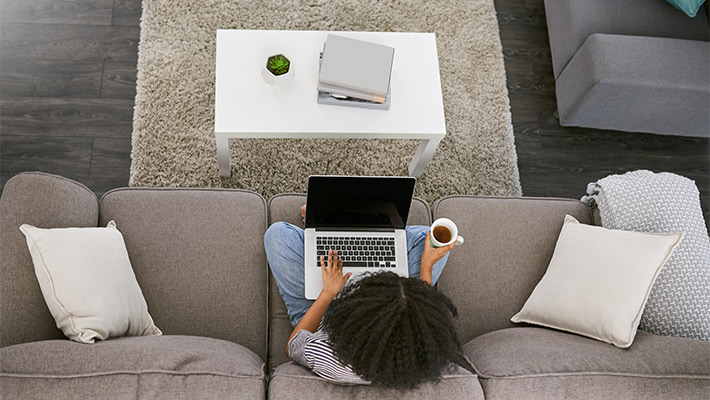 High angle shot of a young woman using a laptop and having coffee on the sofa at home
