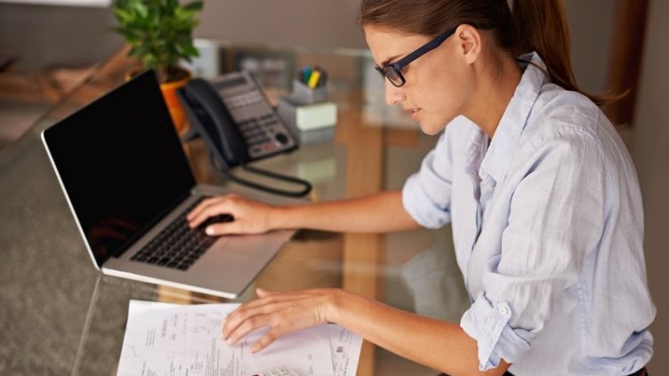 Businesswoman diligently working at her office desk, meticulously reviewing financial documents and receipts while using laptop.