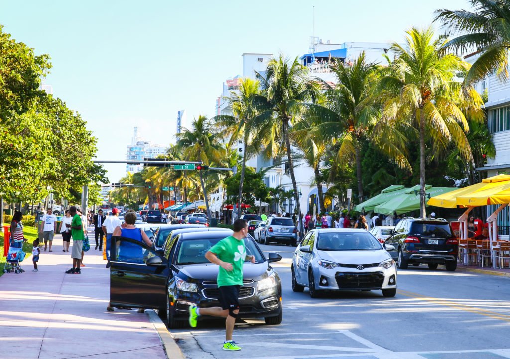 MIAMI BEACH, USA - MAY 9, 2015: Cars driving and parking, people walking by or running, street cafes with sunshades on the side of the Ocean Drive.; Shutterstock ID 395708740